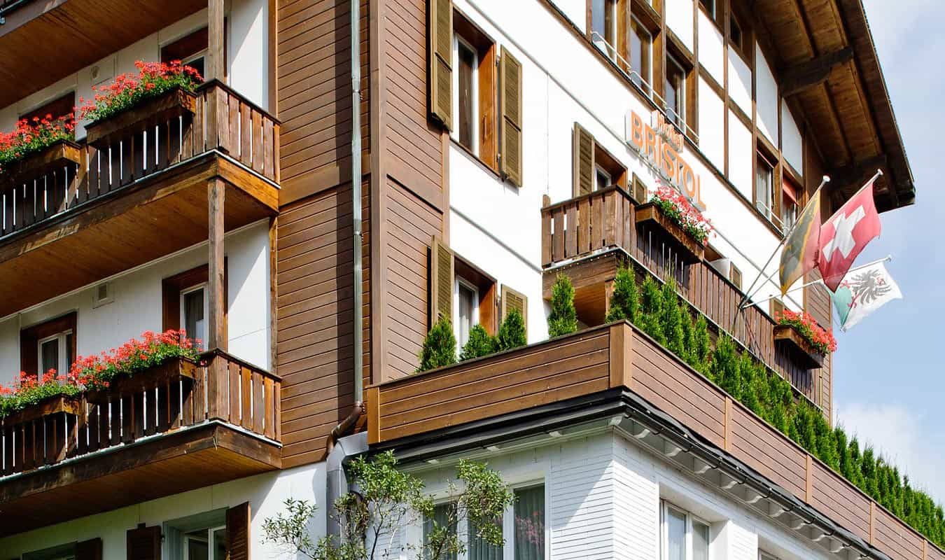Hotel Bristol Adelboden building exterior with wooden balconies, red flowers, and Swiss flags.