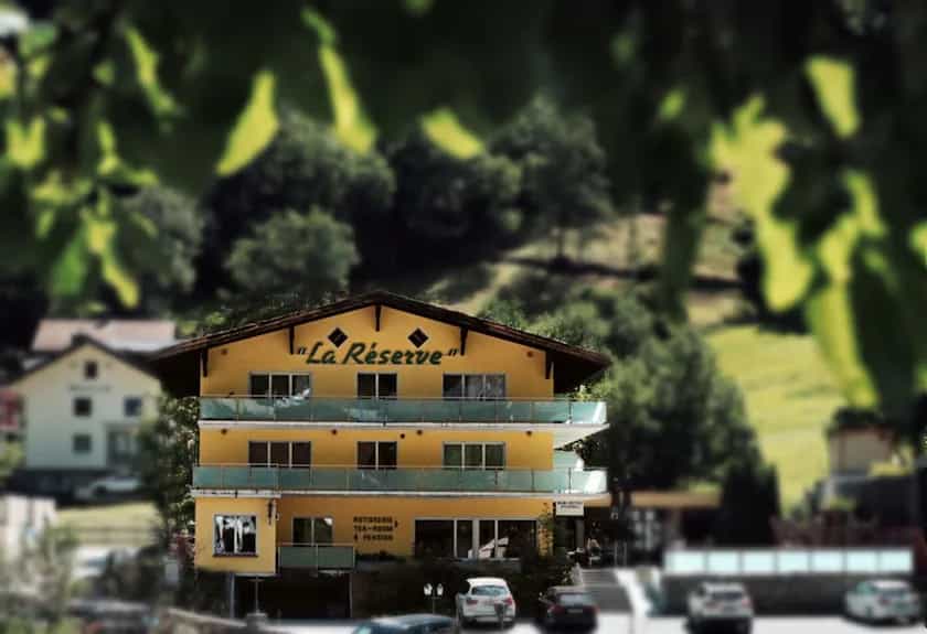Yellow hotel building named "La Réserve" set against a lush green mountain backdrop.