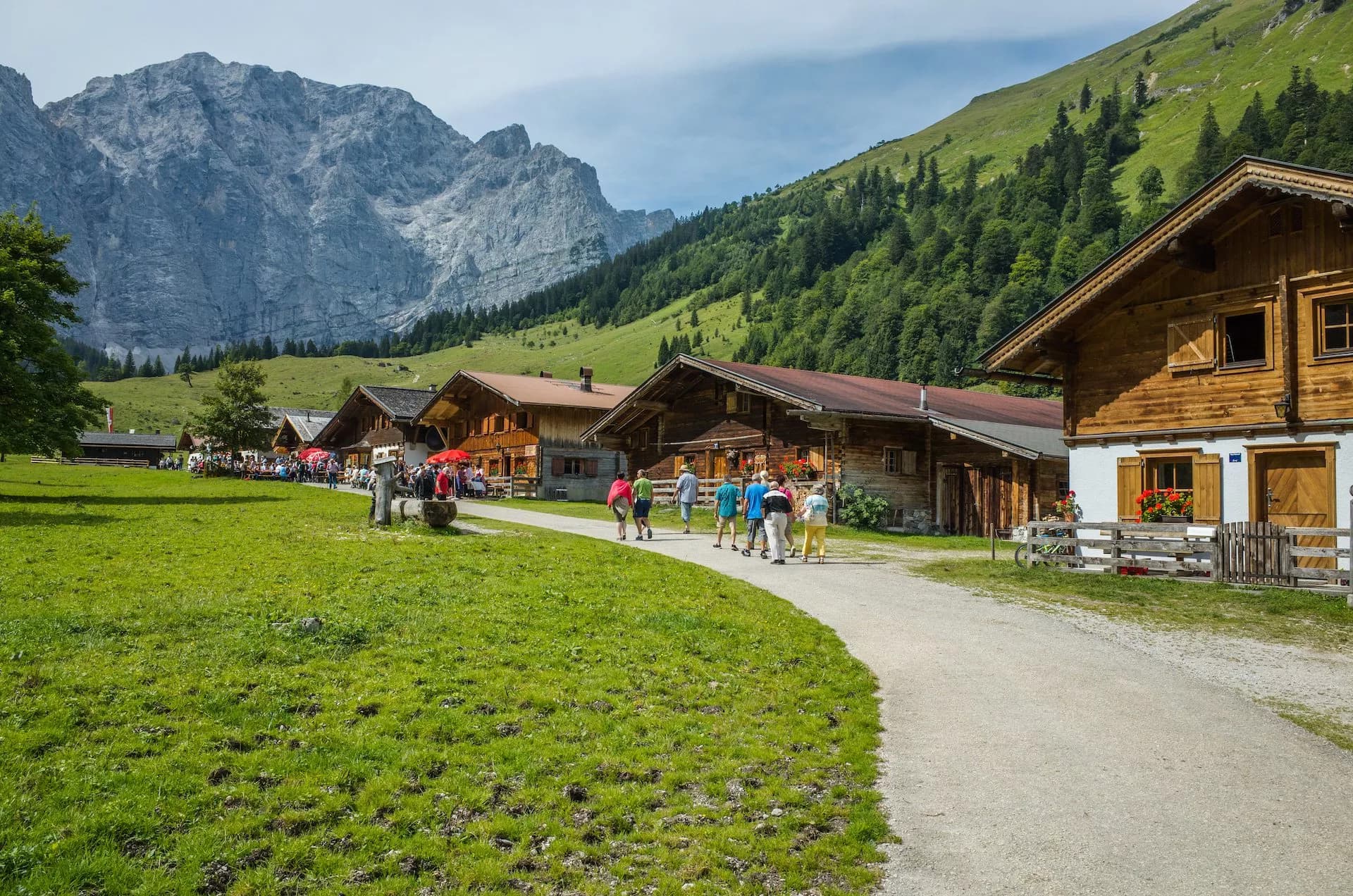Hikers walking past wooden alpine village buildings beneath a large rocky mountain.