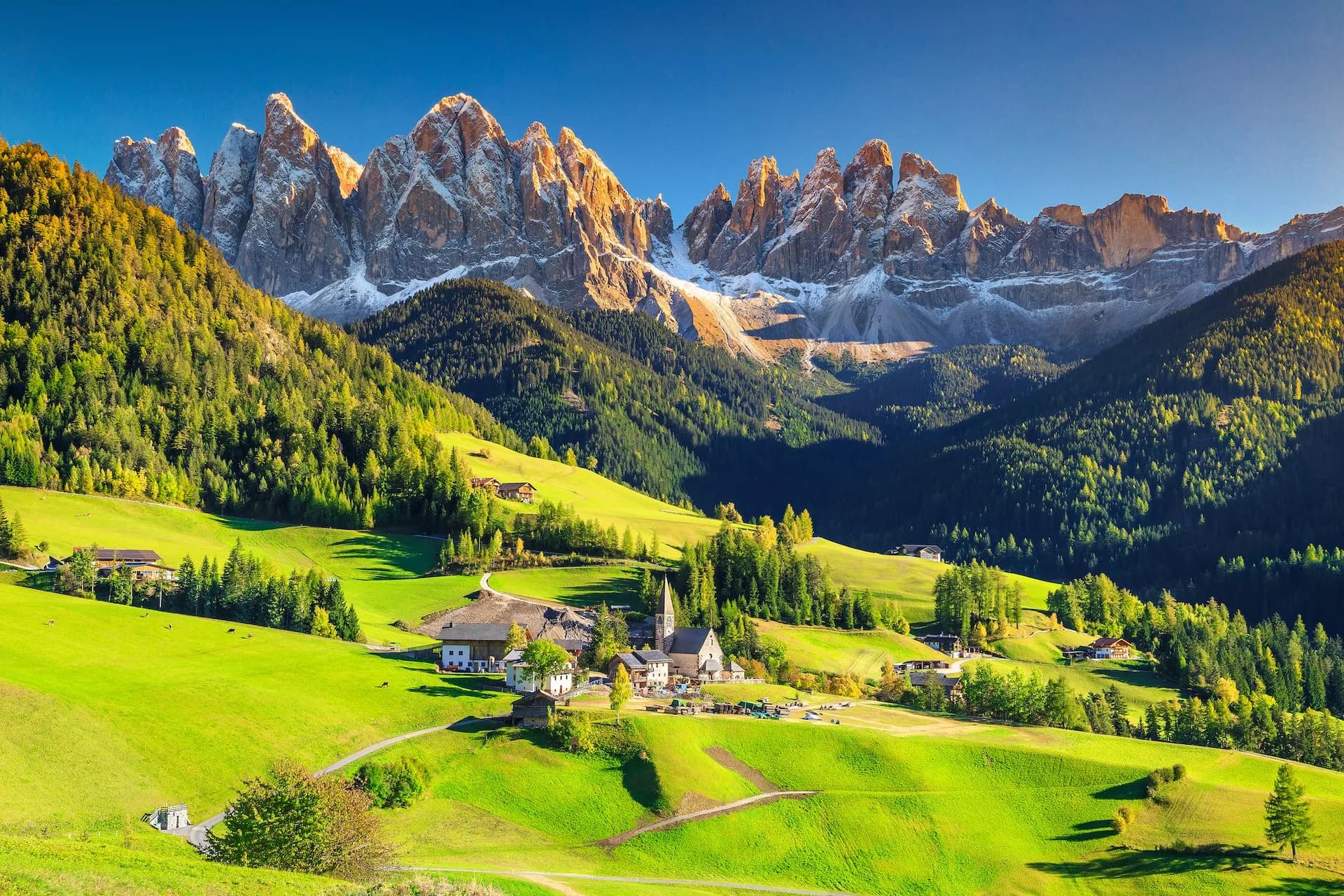 Alpine village with church nestled in green meadows below jagged, sunlit Dolomites mountain peaks