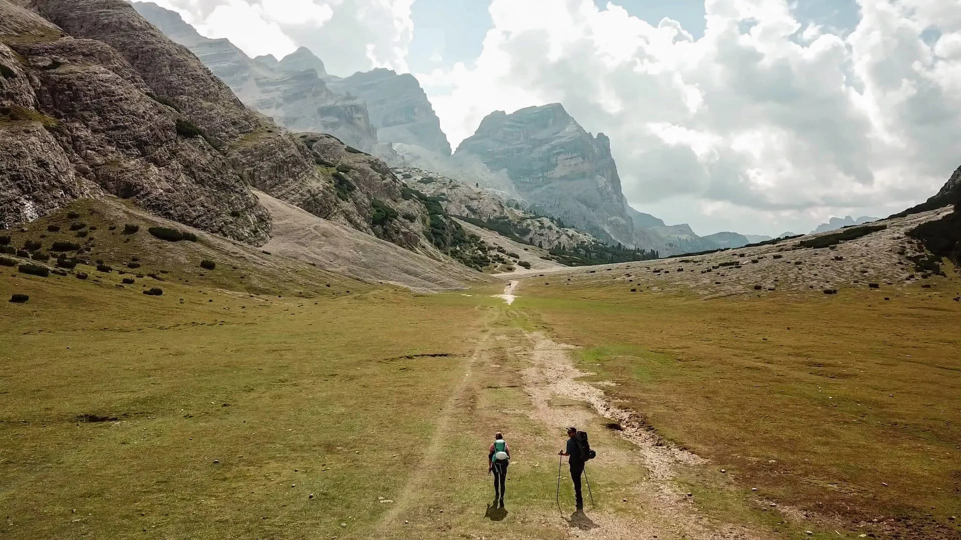 Hikers with backpacks trekking on dirt path through grassy valley toward massive rocky mountains.