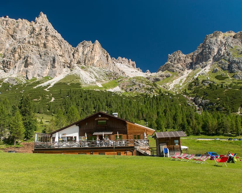 Rifugio Col Gallina mountain hut with outdoor seating below rocky peaks and green forest.