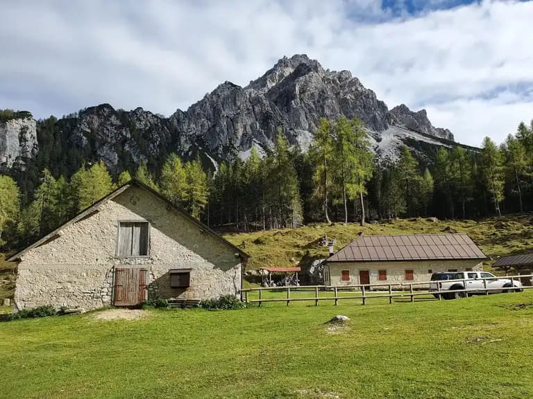 Stone farm buildings in a grassy meadow below a rugged, forested mountain peak, Malga Pramper.