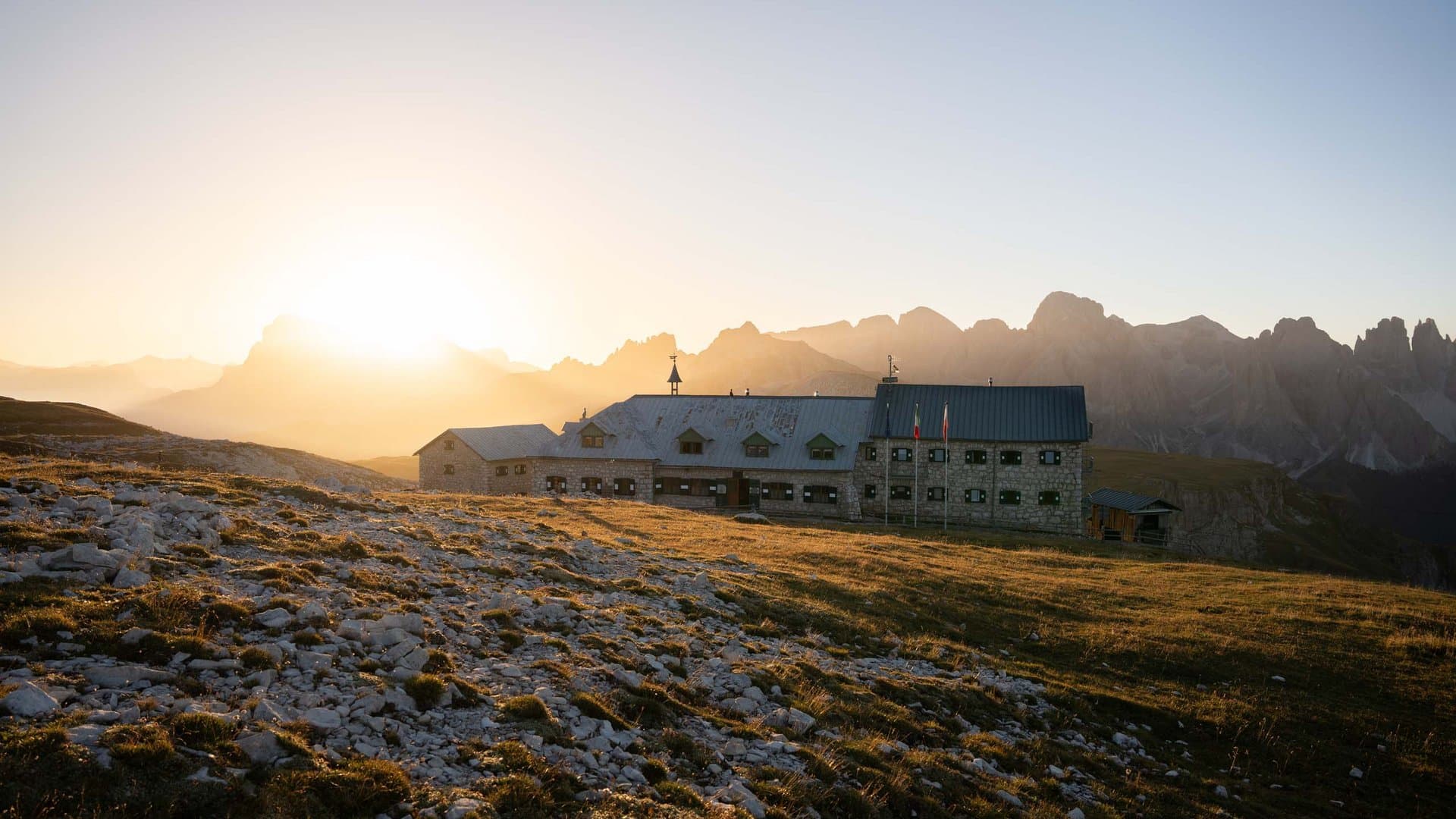 Stone mountain hut in the Dolomites at sunrise with rocky terrain and jagged peaks in the background.