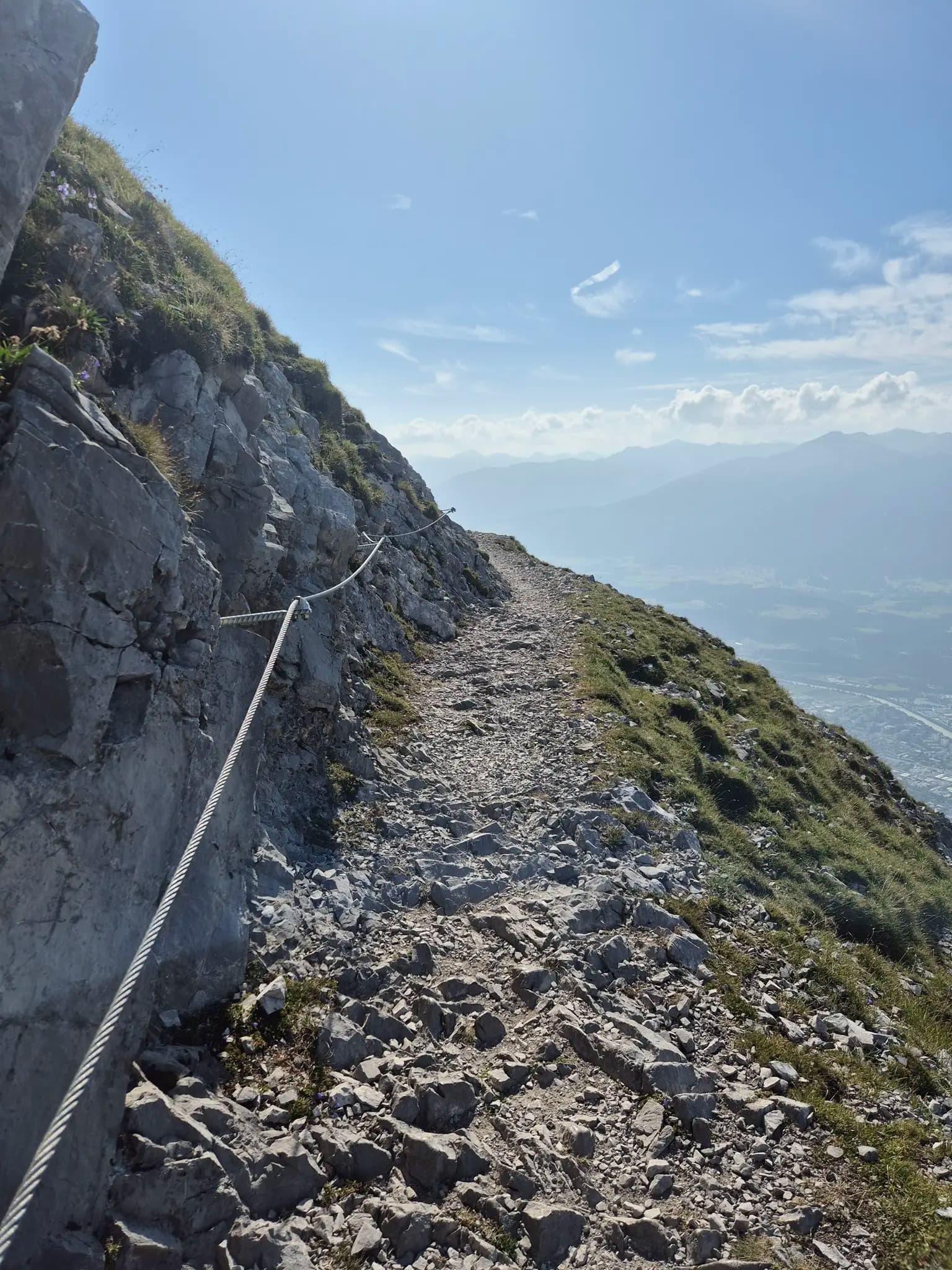Narrow rocky hiking trail with a safety cable along a steep mountain ridge.