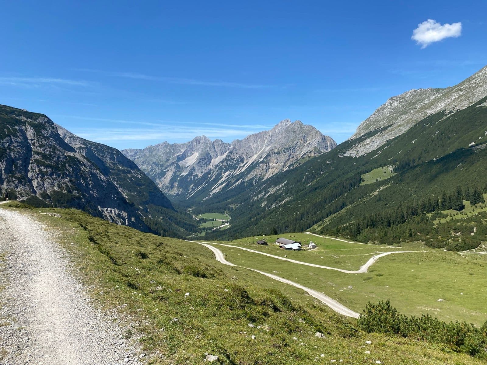 Gravel hiking trail leading into an alpine valley with mountain peaks, green meadows, and a small farm under a clear sky.