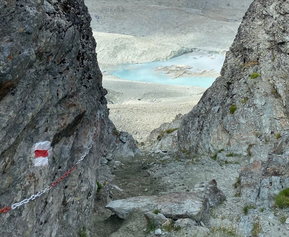 Steep mountain hiking trail with safety chain and red-white trail marker overlooking a glacial lake.