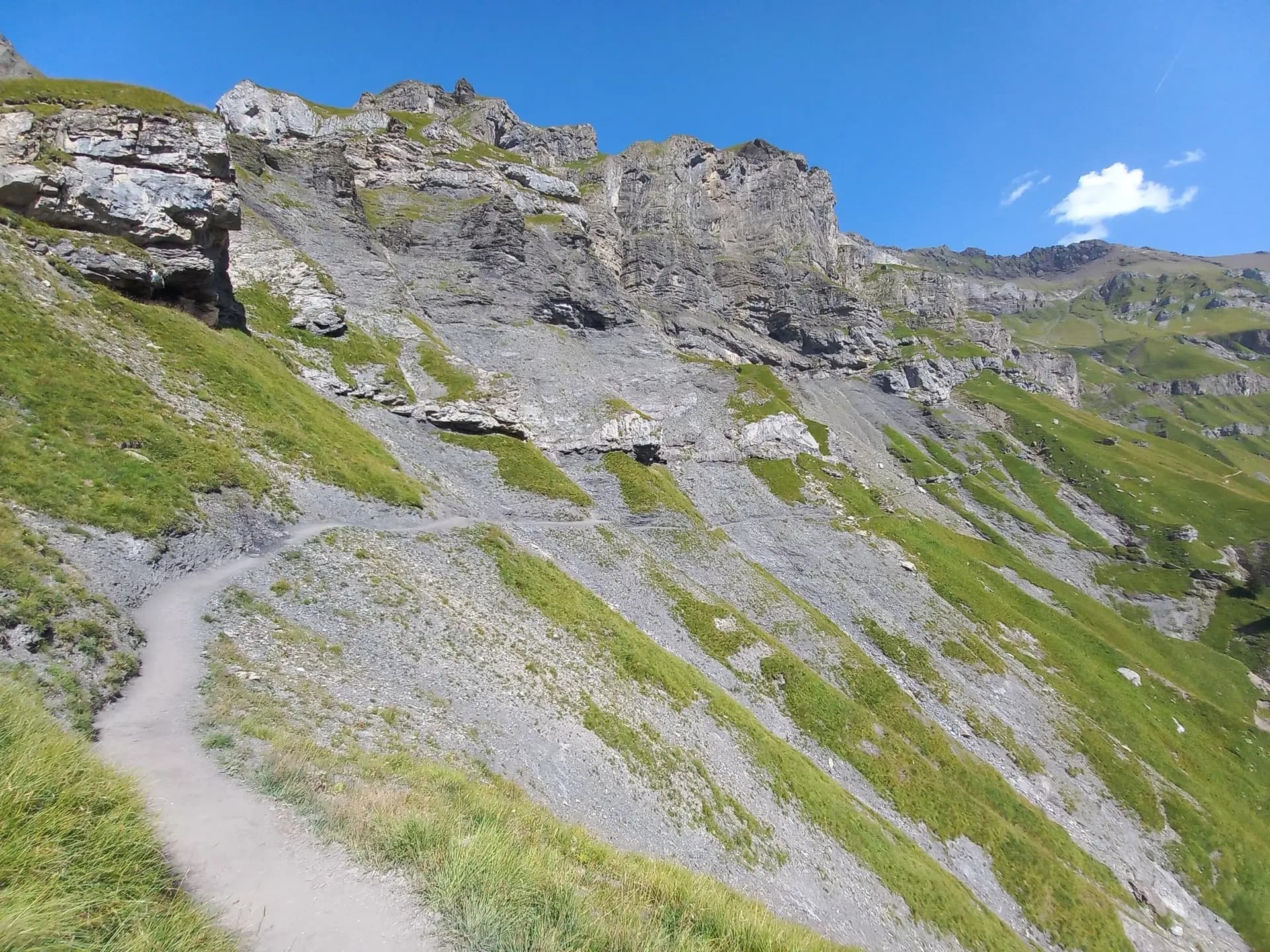 Hiking trail winding along a steep, rocky alpine mountain slope under a clear blue sky.