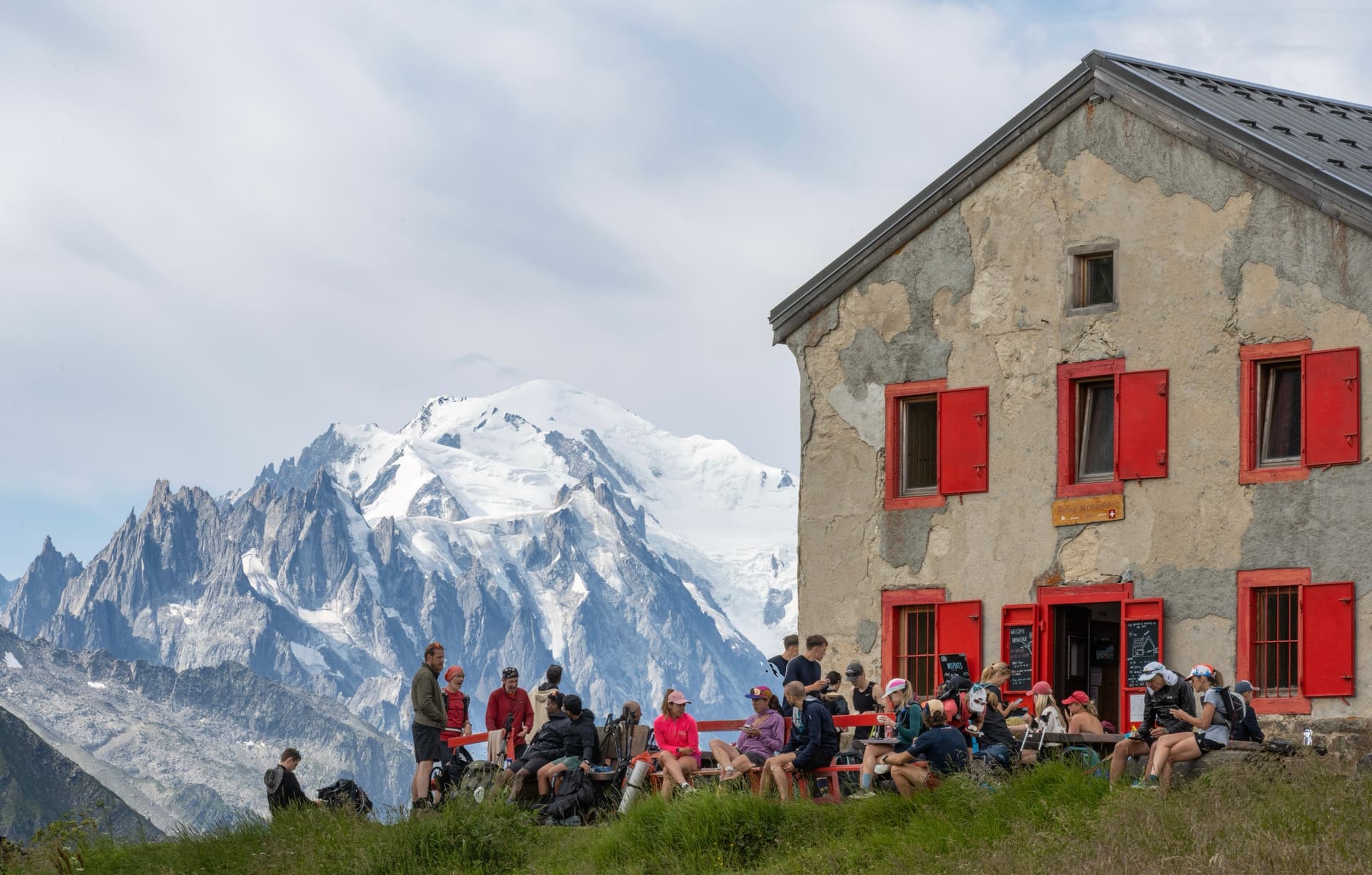 Hikers gathered outside mountain refuge with red shutters in front of snowy peaks