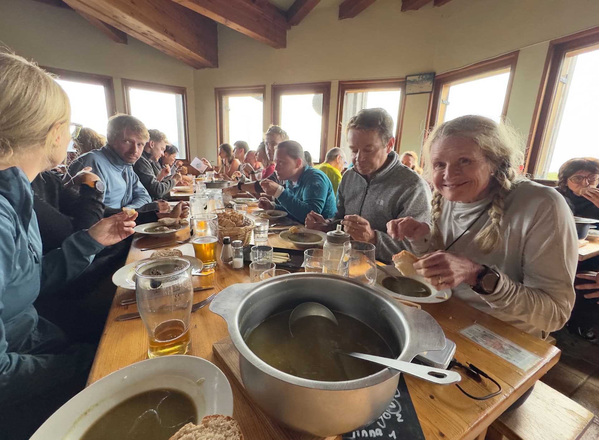 Group dining inside mountain refuge with large pot of soup and drinks on wooden table