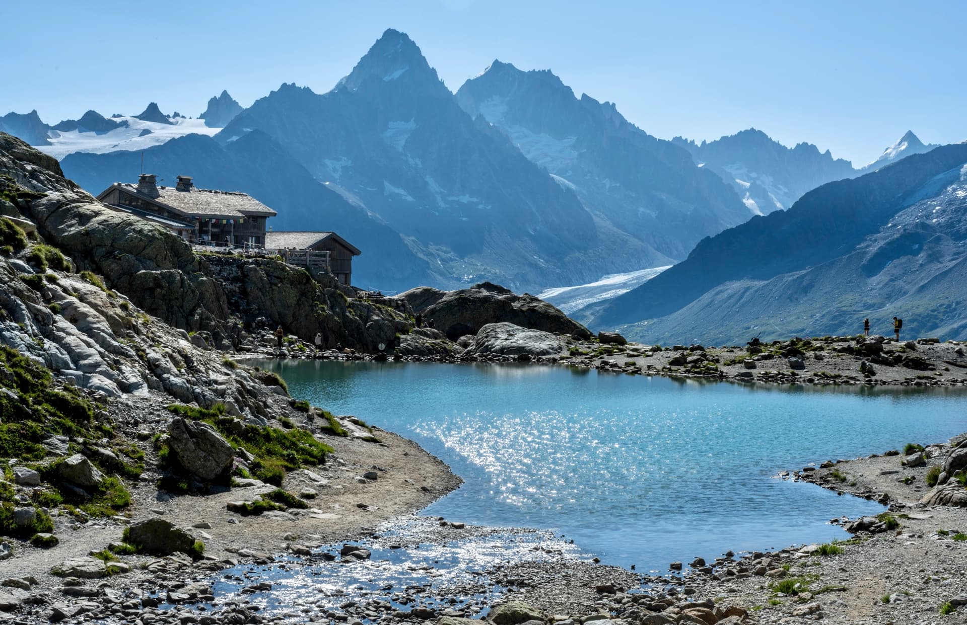 Turquoise alpine lake below rocky mountain peaks with a stone building on the shore
