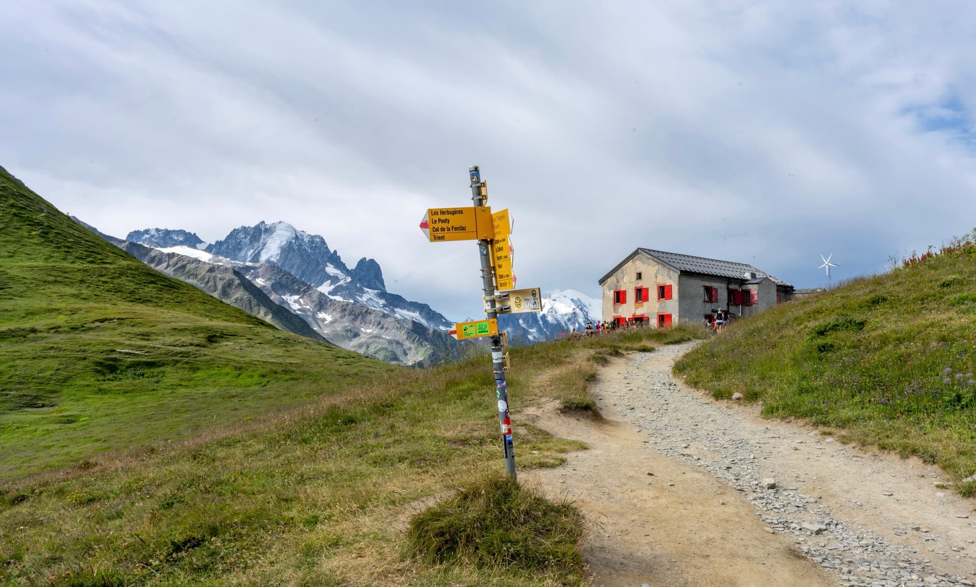 Trail signpost on a mountain hiking path near a stone hut with snowy peaks in the background