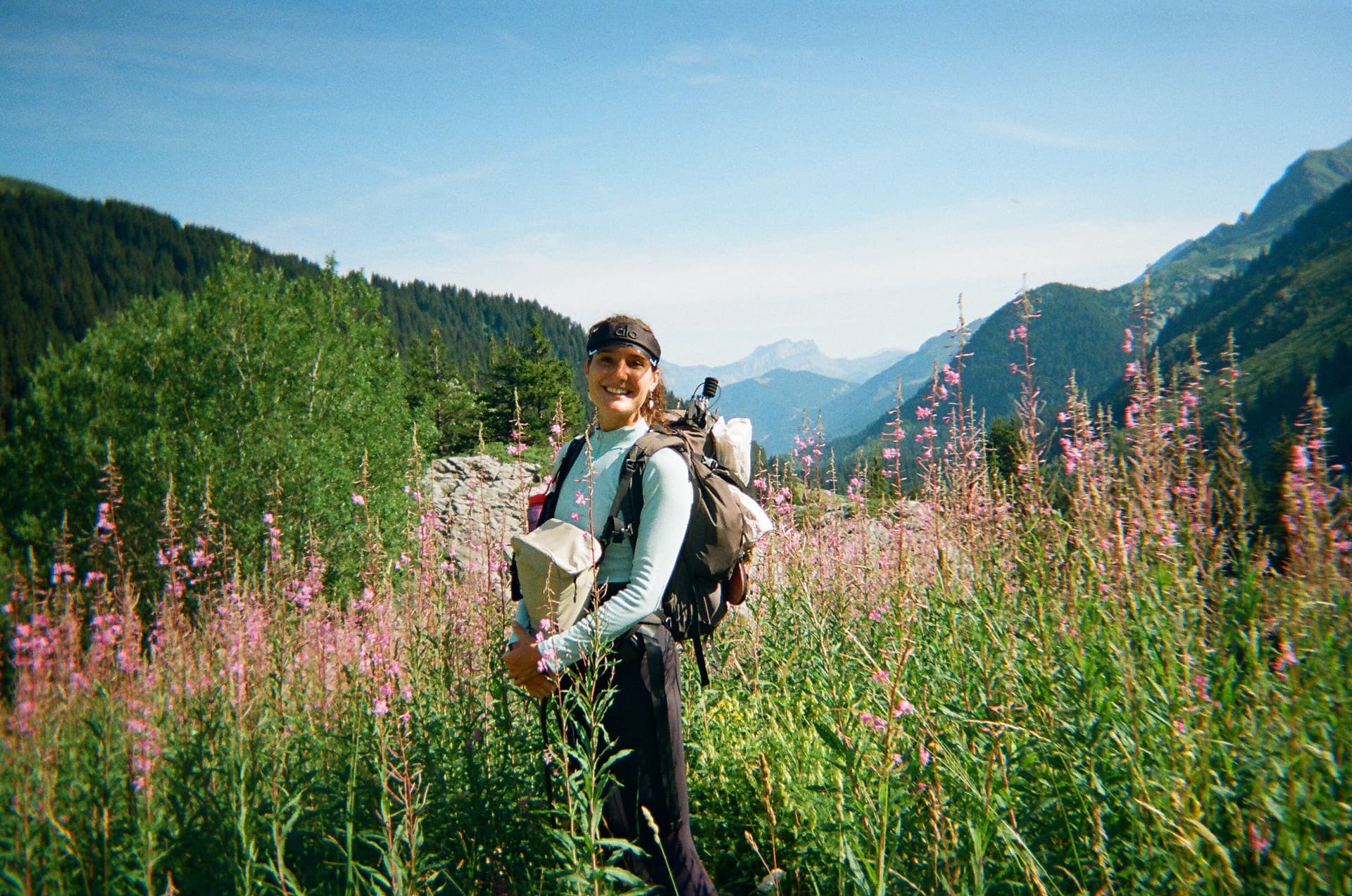 Hiker with backpack standing in a field of pink fireweed wildflowers with mountain and forest background