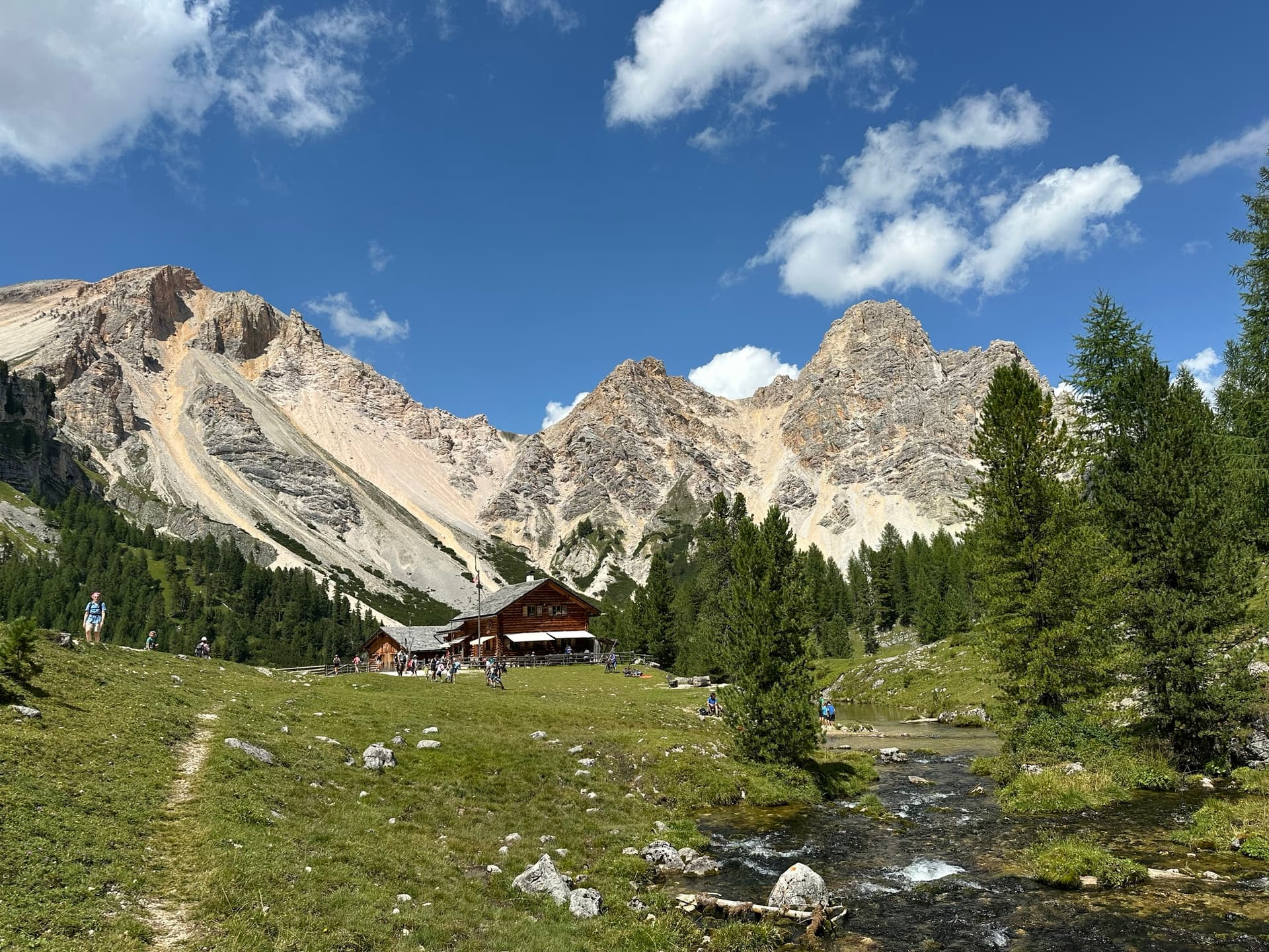 Mountain hut below jagged peaks with stream and hikers in alpine landscape