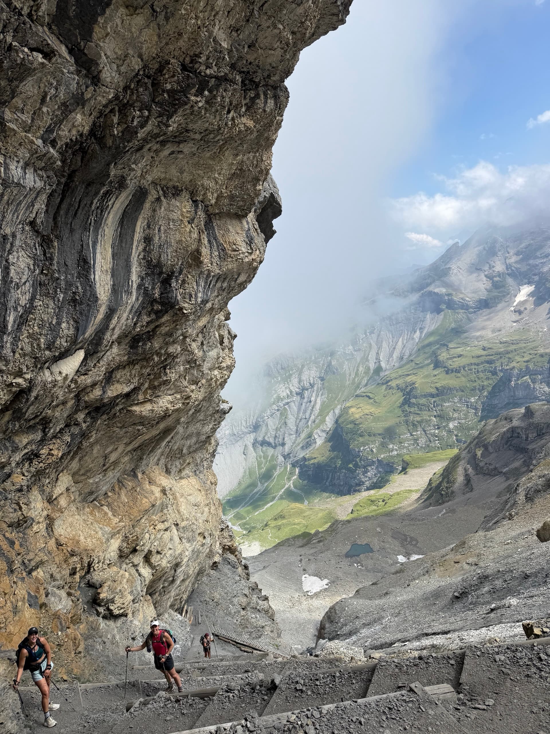 Hikers walking on a mountain trail beneath an overhanging rock wall with valley views