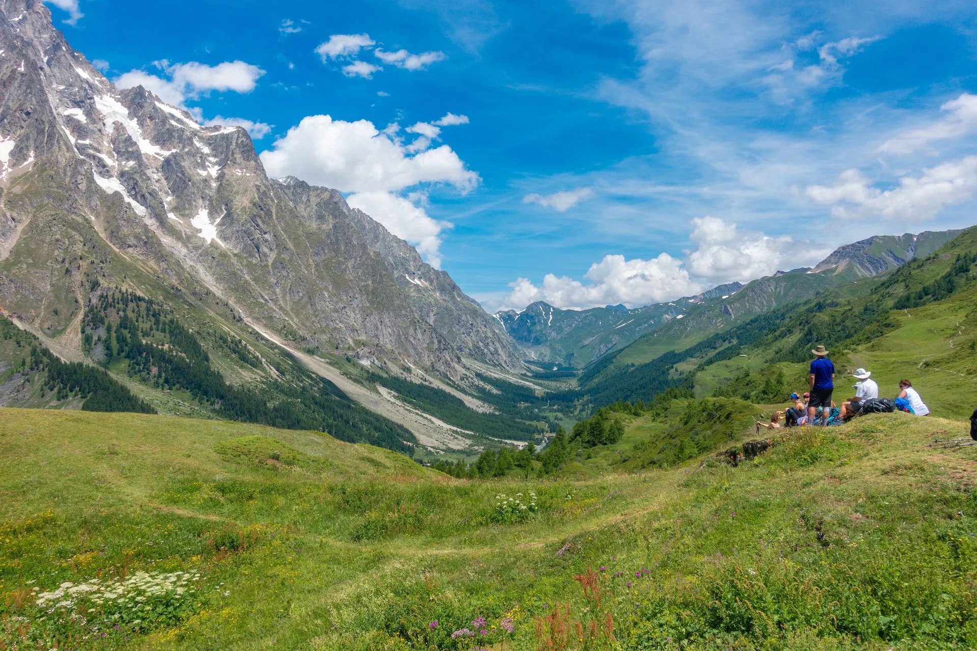 Hikers resting overlooking Val Ferret alpine valley with snow-capped mountains and green slopes.