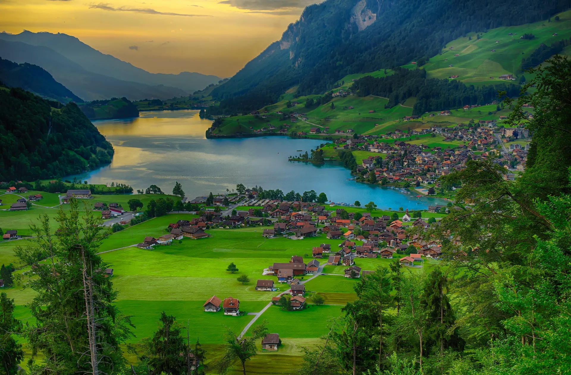 Alpine village on green valley beside bright blue lake with steep forested mountains at sunset