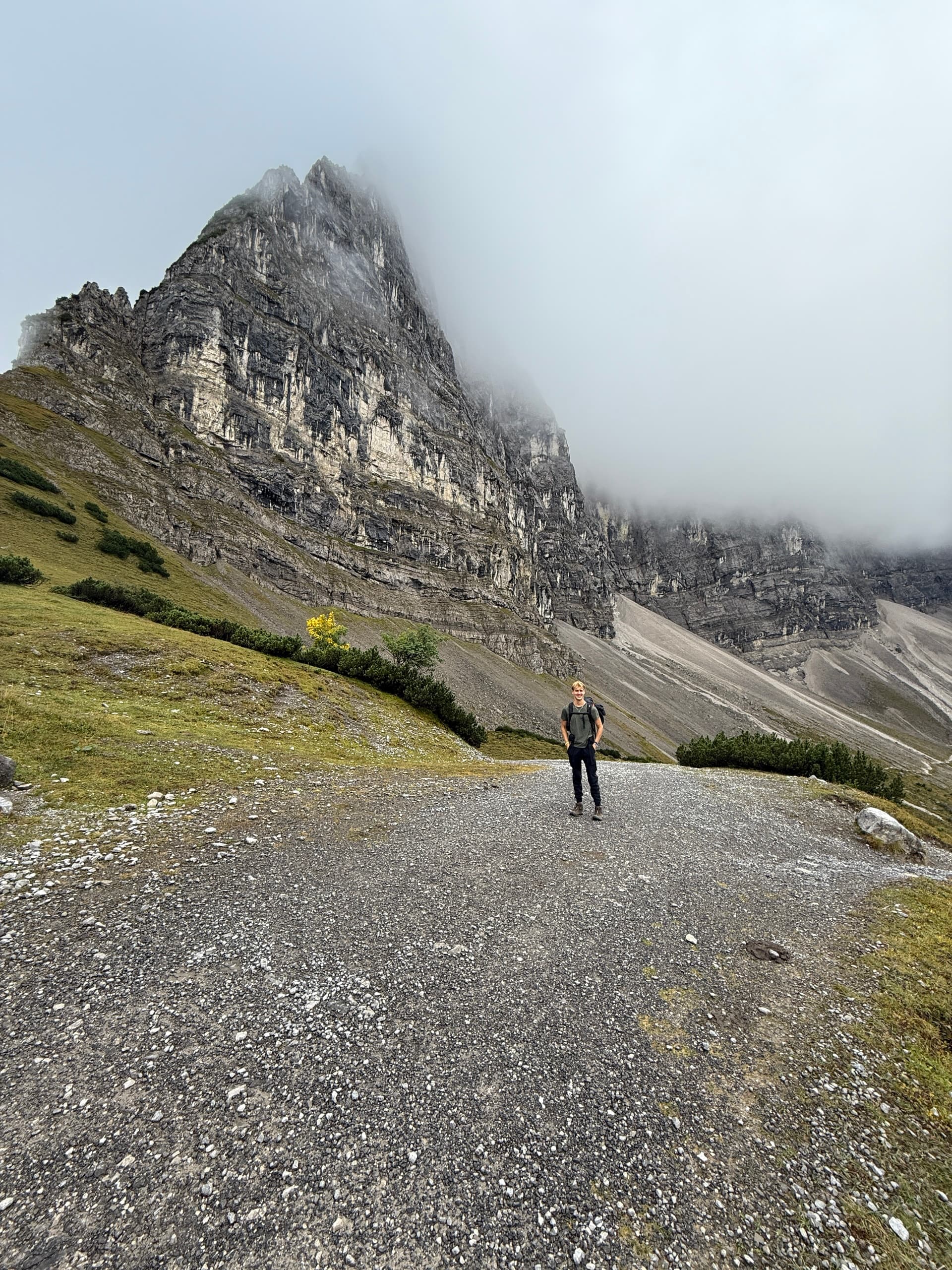 Hiker standing on gravel path below rocky mountain peak shrouded in mist